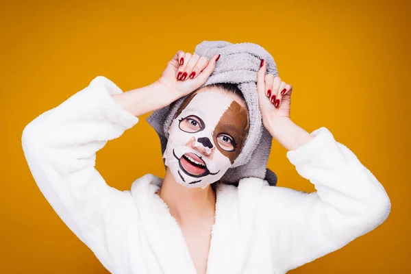 Vrolijke jonge meisje met een witte handdoek op haar hoofd is plezier, op haar gezicht een hydraterende masker met een dog's gezicht — Stockfoto