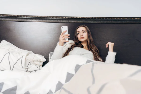 Triste niña somnolienta se encuentra en la cama bajo una manta blanca, sostiene un teléfono inteligente y quiere dormir — Foto de Stock