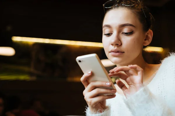 Serious young girl in a white warm sweater writes a message on her smartphone — Stock Photo, Image