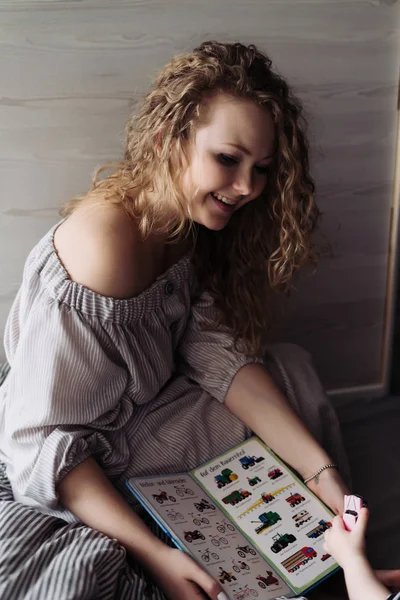 A young curly woman in a beautiful dress teaches her little son to read, laughs — Stock Photo, Image