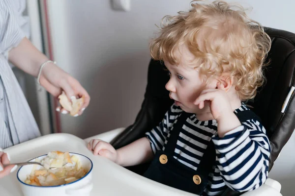 Маленький кудрявый мальчик сидит на кухне, мать кормит вкусный горячий суп — стоковое фото