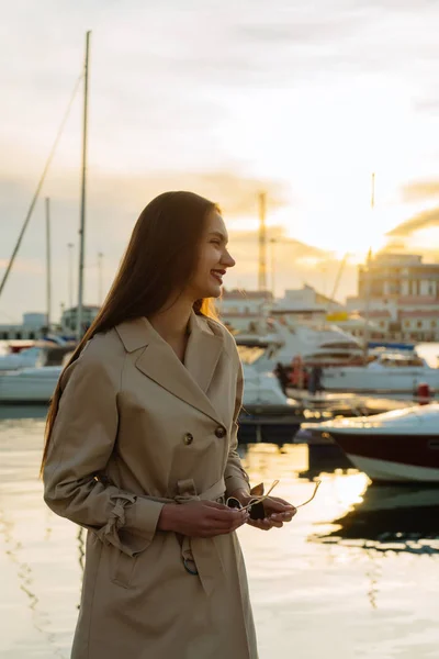 Elegante ragazza dai capelli lunghi in un cappotto beige alla moda in attesa di uno yacht nel porto, ridendo — Foto Stock