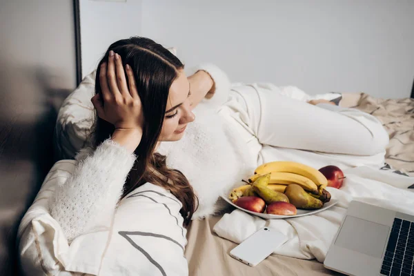 흰 잠 옷에서 여자 침대, 휴식 하 고, 그녀의 노트북에 재미 있는 영화를 보고 하 고 먹는 과일에에서 속 인 다 — 스톡 사진