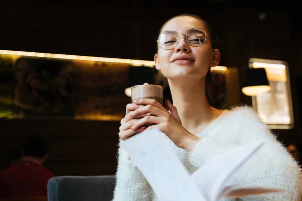 Чарівна молода жінка в окулярах і білий светр п'є ароматний латте в кафе, посміхаючись — стокове фото