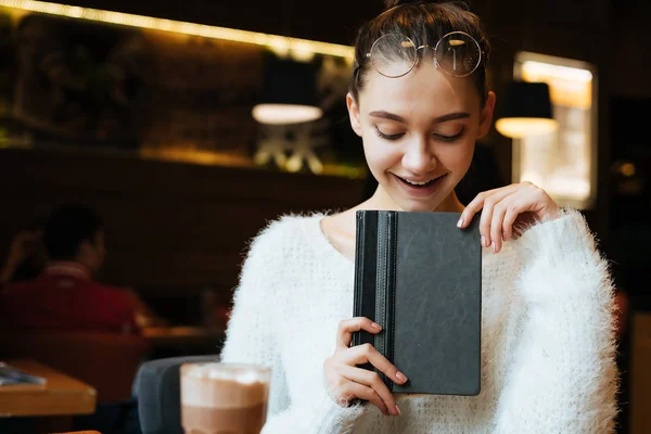 Очаровательная девушка фрилансер в белом свитере сидит в кафе и отдыхает, в руках черный блокнот — стоковое фото