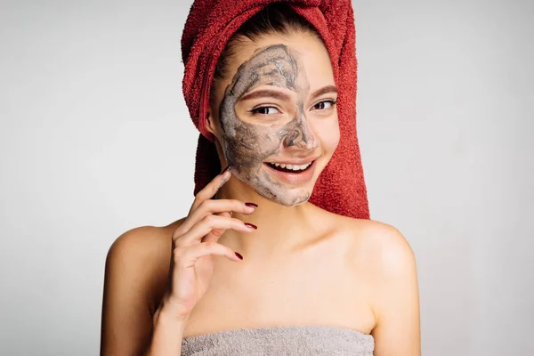 Μια γοητευτική νεαρή κοπέλα με μια κόκκινη πετσέτα στο κεφάλι της ένα χρήσιμο πήλινο προσωπείο εφαρμόζεται στο μισό πρόσωπο — Φωτογραφία Αρχείου