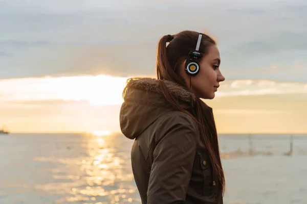Ung vacker flicka i en varm jacka som promenader vid havet i solnedgången, lyssna på musik i hörlurar — Stockfoto