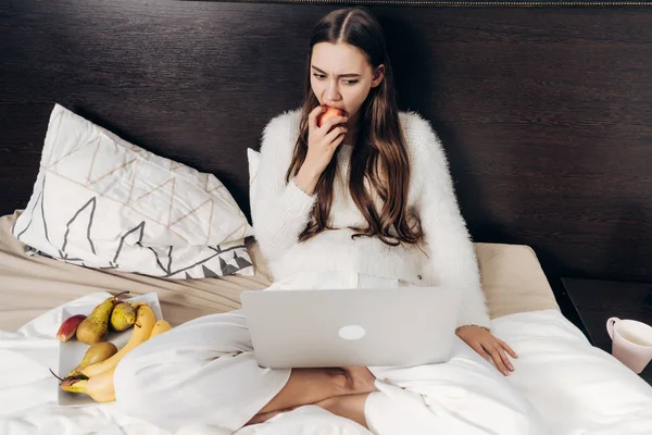 Молода дівчина з довгим волоссям, сидячи в ліжку, дивиться телевізійне шоу на її ноутбук і їсть яблуко — стокове фото