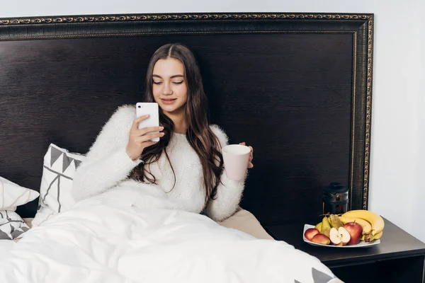 Schattig slaperig meisje met lang haar in bed ligt, houdt van smartphone, wil slapen — Stockfoto