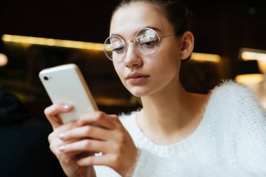 ciddi genç kız öğrenci gözlük ve beyaz bir kazak onun smartphone görünüyor, okuldan sonra bir kafede oturuyor