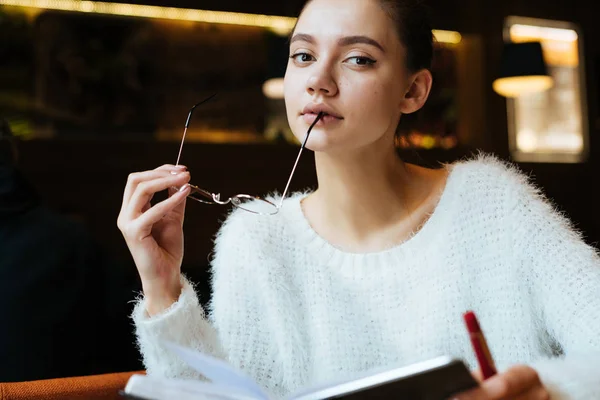 Серьезная молодая девушка фрилансер в белом свитере сидит в кафе, думая о работе — стоковое фото
