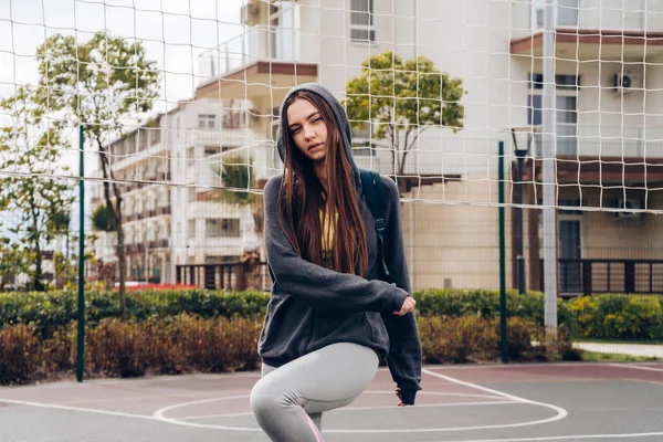 Активна довгошерста дівчина в сірій куртці міняє м'язи перед тренуванням на спортивному майданчику — стокове фото