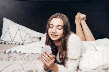 Beyaz pijamalı güzel uzun saçlı kız yatakta yatıyor, onu smartphone ve gülümser bir ileti yazar