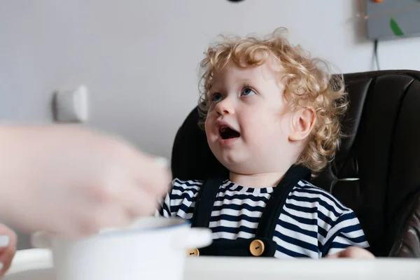 Rubio rizado bebé niño sentado en la cocina, mamá alimentación deliciosa comida, desayuno familiar — Foto de Stock