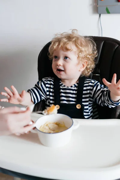 Счастливый маленький кудрявый мальчик с голубыми глазами, сидящий на кухне и кушающий вкусную еду вместе с мамой — стоковое фото