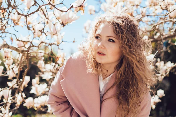 Elegant curly woman in a pink coat walks in the park next to the blooming magnolia, a good mood — Stock Photo, Image