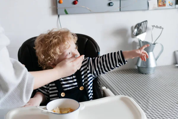 Маленький светловолосый мальчик ест вкусную еду на кухне, заботливая мать вытирает ему рот — стоковое фото