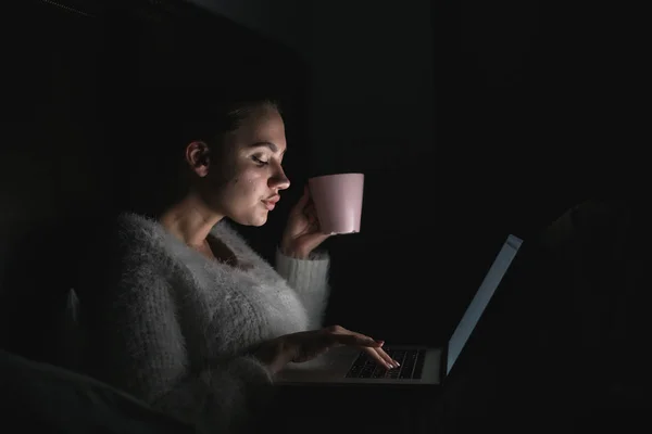 Молода дівчина фрілансер працює на ноутбуці пізно вночі, п'є каву — стокове фото