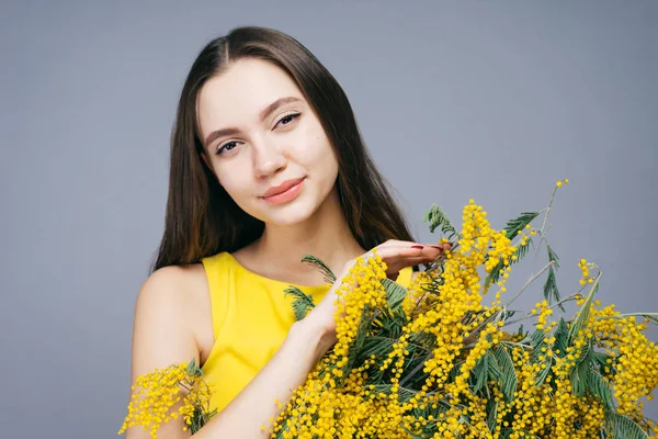 Encantadora chica hermosa en un vestido amarillo de moda está sosteniendo una mimosa fragante en las manos, mirando a la cámara — Foto de Stock
