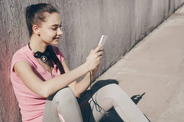 強さを増しているトレーニングと彼女のスマート フォンを検討する前に、太陽の下で地面に座って美しい少女の笑みを浮かべてください。 — ストック写真