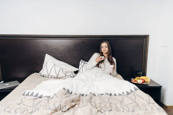 Jong meisje zit in bed onder een witte deken, moe en wil slapen, heeft een smartphone in haar handen — Stockfoto