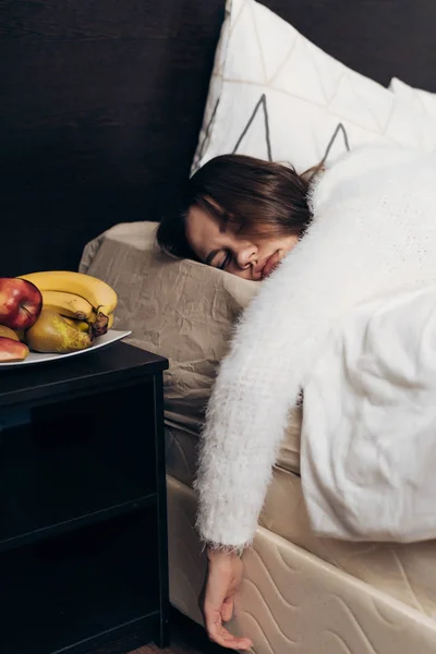 Een jong meisje slaapt op een bed onder een witte deken, wil niet opstaan, naast haar zijn vrucht — Stockfoto