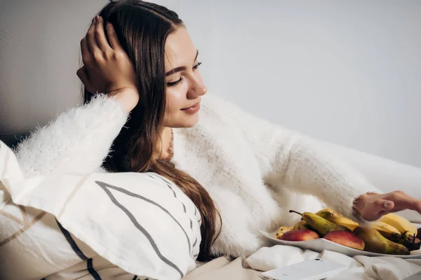 Una chica dulce y somnolienta yace en una cama en pijama blanco, descansando y comiendo fruta — Foto de Stock