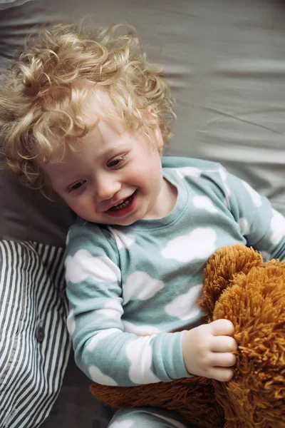 Pequeño niño de pelo rubio en lindo pijama azul se ríe, sostiene su juguete relleno — Foto de Stock