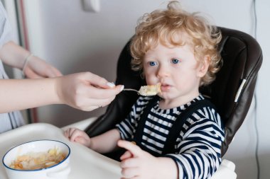 sarı saçlı mavi gözlü küçük çocukla mutfağa yüksek sandalyede oturuyor, annem yararlı yulaf lapası beslemek