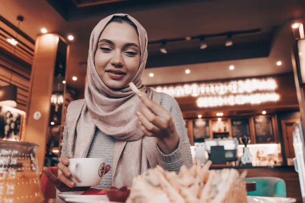 Schöne arabische Mädchen mit Kopftuch auf dem Kopf essen leckeres warmes Essen in einem Café, lächelnd — Stockfoto