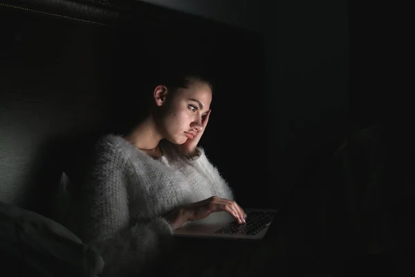 Уставшая молодая девушка сидит в постели поздно ночью в темноте и смотрит скучный фильм на своем ноутбуке — стоковое фото