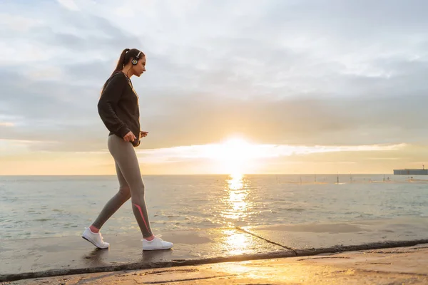 Ragazza atletica in abbigliamento sportivo a riposo dopo aver fatto jogging al mare al tramonto, ascoltando musica sulle cuffie — Foto Stock