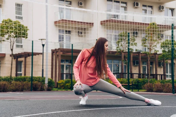 Chica delgada y flexible en una chaqueta rosa se estira y calienta los músculos en el campo de deportes, lleva un estilo de vida activo — Foto de Stock