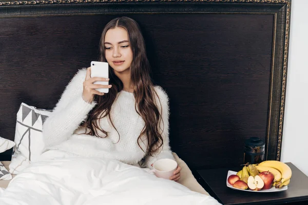 Hermosa chica con el pelo largo se sienta en la cama por la noche, cansado y quiere dormir, se enciende el despertador en su teléfono inteligente — Foto de Stock