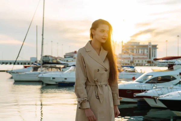 En ung omtänksam tjej med långt hår, i en beige mode kappa väntar på sin yacht i hamnen — Stockfoto