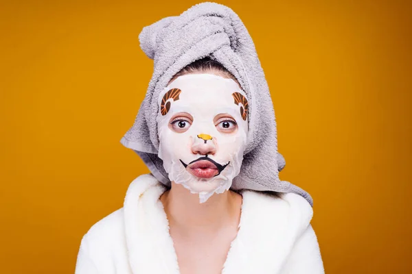 Une jeune fille surprise en robe de chambre éponge blanche, avec une serviette sur la tête profitant du spa, sur son visage un masque hydratant avec un motif — Photo