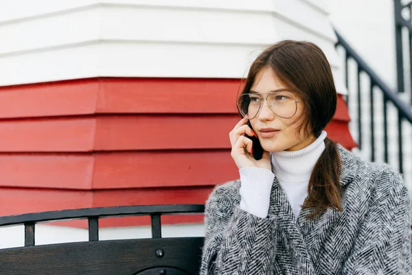 Мила молода брюнетка дівчина в окулярах і сіре пальто розмовляє по телефону, сидячи на лавці на відкритому повітрі, вуличному стилі і моді — стокове фото