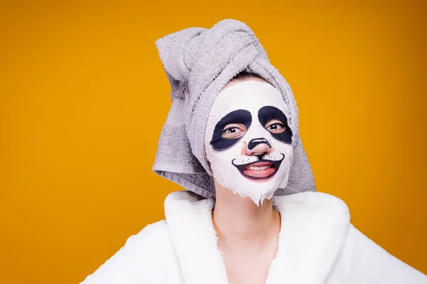 girl in a white robe and with a towel on her head smiling and posing on a yellow background, on her face a moisturizing mask with a panda face