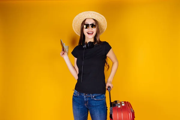 Счастливая девушка в соломенной шляпе и солнцезащитных очках идет в школу с большим красным чемоданом, ждет свой самолет — стоковое фото