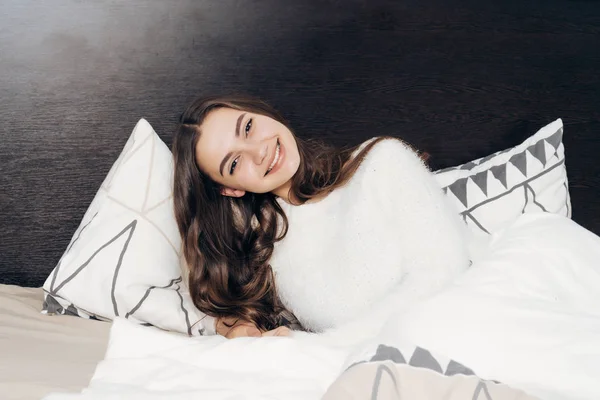 Счастливая длинноволосая девушка в белой пижаме ложится рано утром в постель, в хорошем настроении и улыбается — стоковое фото