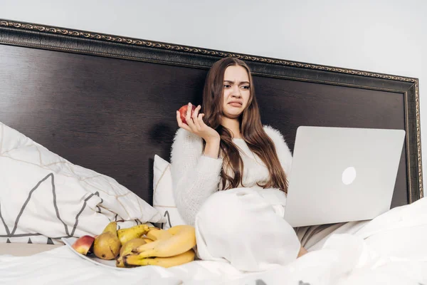 Une jeune fille mécontente est assise dans son lit sous une couverture blanche, regarde quelque chose de désagréable sur son ordinateur portable et mange une pomme — Photo