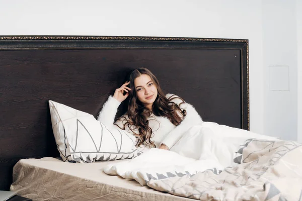 Sonolento menina de cabelos longos em pijama branco fica na cama, descansando depois de um dia de trabalho duro — Fotografia de Stock