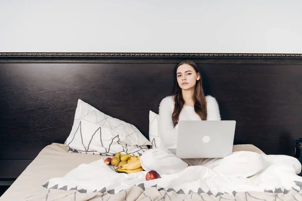 Zmęczony młoda dziewczyna w piżamie biały siedzi w łóżku wieczorem, pracy na jej laptopie i jedzenie owoce — Zdjęcie stockowe