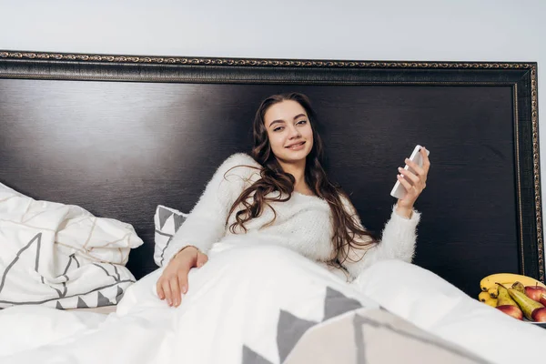 Όμορφο κορίτσι υπνηλία έγκειται στο κρεβάτι κάτω από μια άσπρη κουβέρτα, ξύπνησα το πρωί σε μια καλή διάθεση, κρατά ένα smartphone — Φωτογραφία Αρχείου