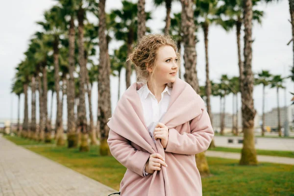 Elegant young woman in a pink coat walking through a green park, enjoying the spring and smiling — Stock Photo, Image