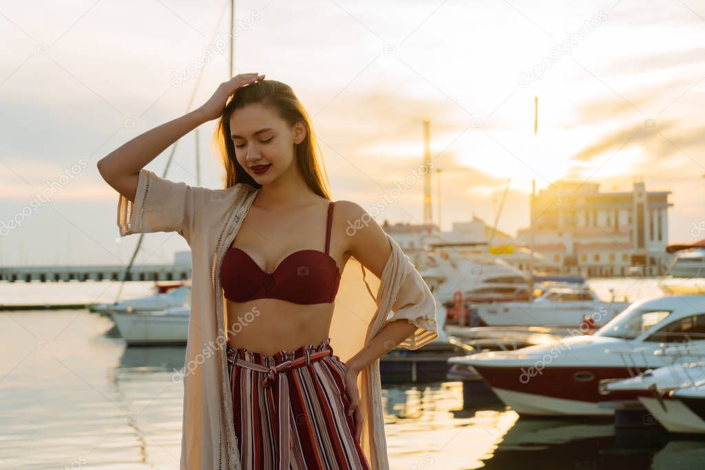 luxurious girl in stylish summer clothes with long hair posing in seaport at sunset