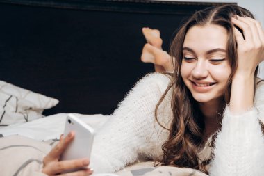 uzun saçlı güzel kız sabah erken uyandım, yatakta yatıyor ve onun smartphone, gülümser bir ileti yazar