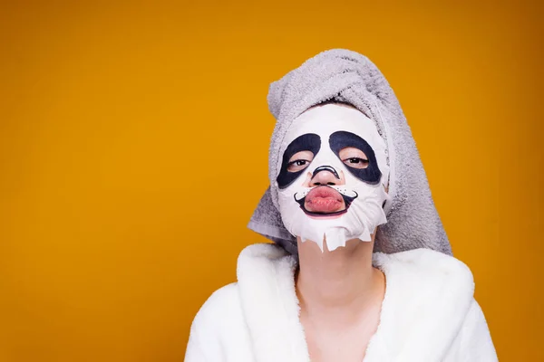 Ένα αστείο κορίτσι με μια ρόμπα terry και με μια πετσέτα στο κεφάλι της που φαίνεται στην κάμερα, στο πρόσωπό της μια ενυδατική μάσκα με πρόσωπο panda — Φωτογραφία Αρχείου