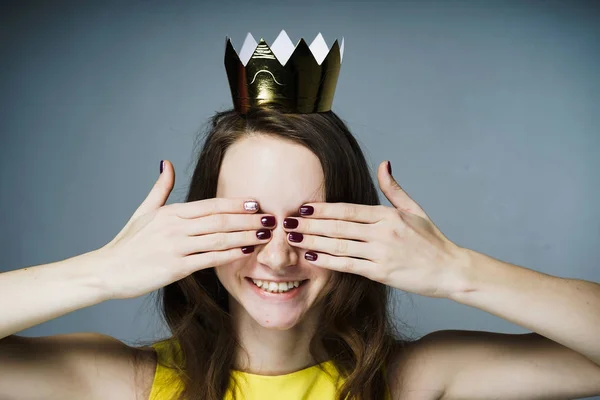 Una linda chica sonriente en un vestido amarillo cubrió sus ojos con sus manos, una corona de oro en su cabeza — Foto de Stock