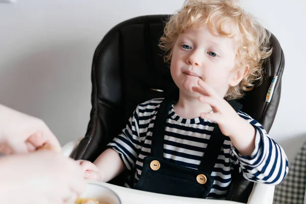 Маленький кудрявый мальчик сидит на кухне на стуле, ест здоровую кашу — стоковое фото